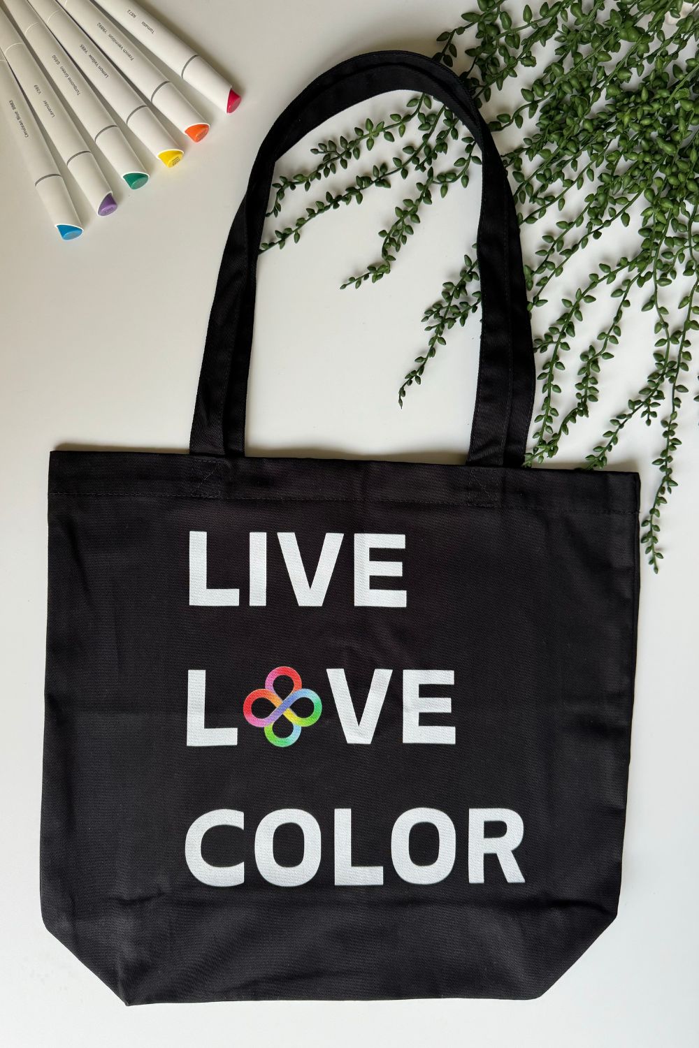 Black canvas tote bag with white bold print that says LIVE LOVE COLOR. The letter O in the word LOVE has been replaced with rainbow infinity symbols.