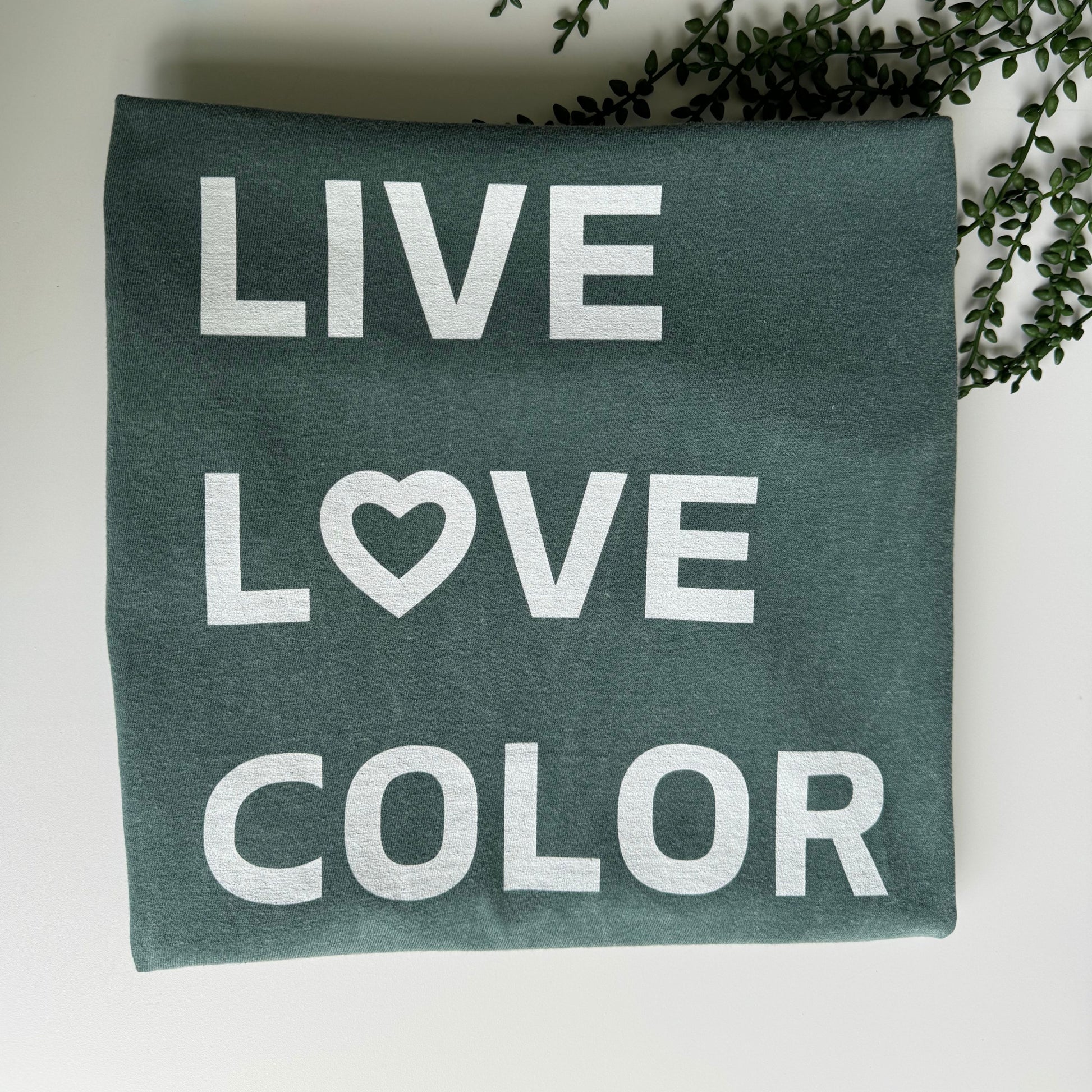 Blue Spruce color T-Shirt with white bold print that says LIVE LOVE COLOR. The letter O in the word LOVE has been replaced with a heart symbol.