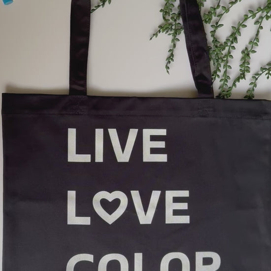 Black canvas tote bag with white bold print that says LIVE LOVE COLOR. The letter O in the word LOVE has been replaced with a heart symbol.