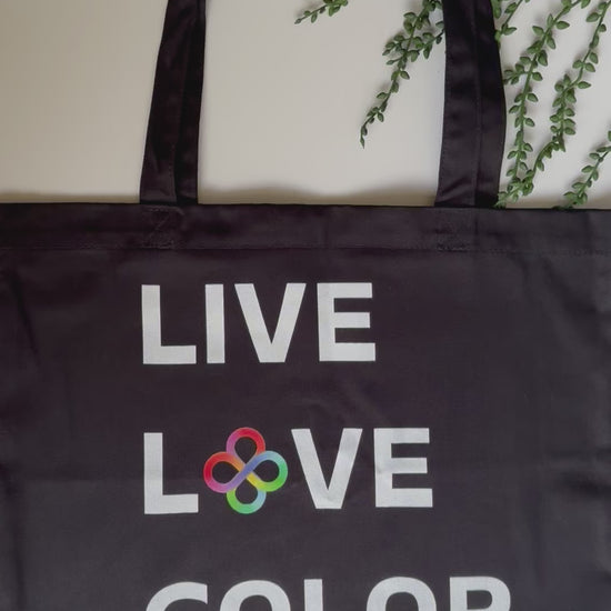 Black canvas tote bag with white bold print that says LIVE LOVE COLOR. The letter O in the word LOVE has been replaced with rainbow infinity symbols.